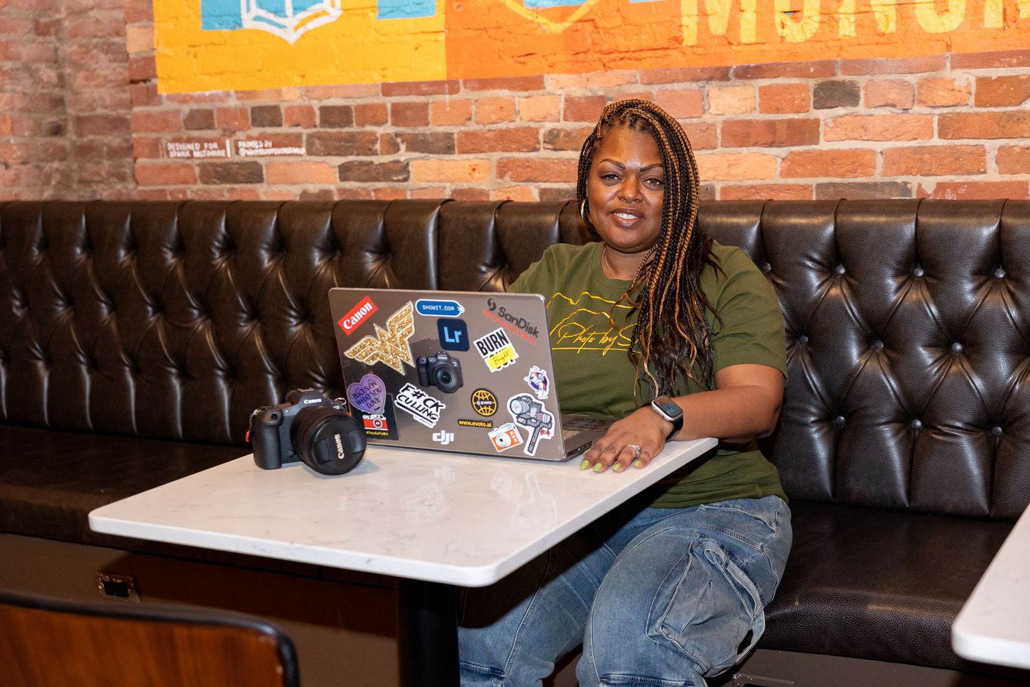 Woman in olive shirt sitting at table with laptop and camera in brick interior.