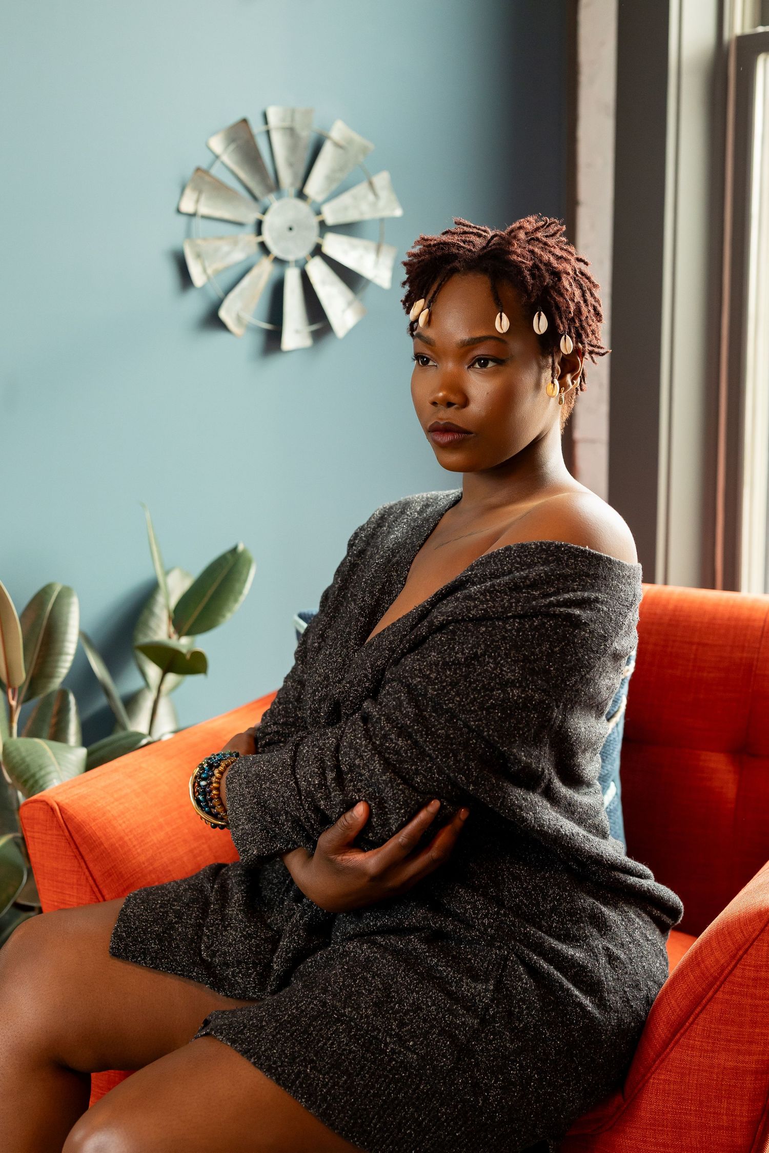Woman with short braided hair sitting in orange chair wearing gray off-shoulder dress.