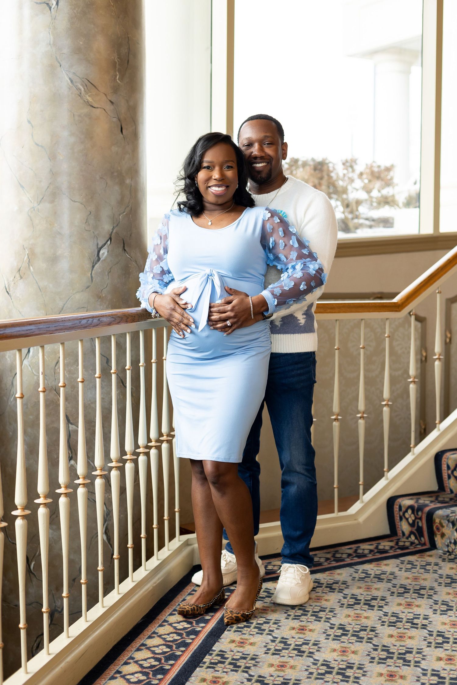 Pregnant woman in blue dress and man in white sweater pose together on elegant staircase for maternity portrait.