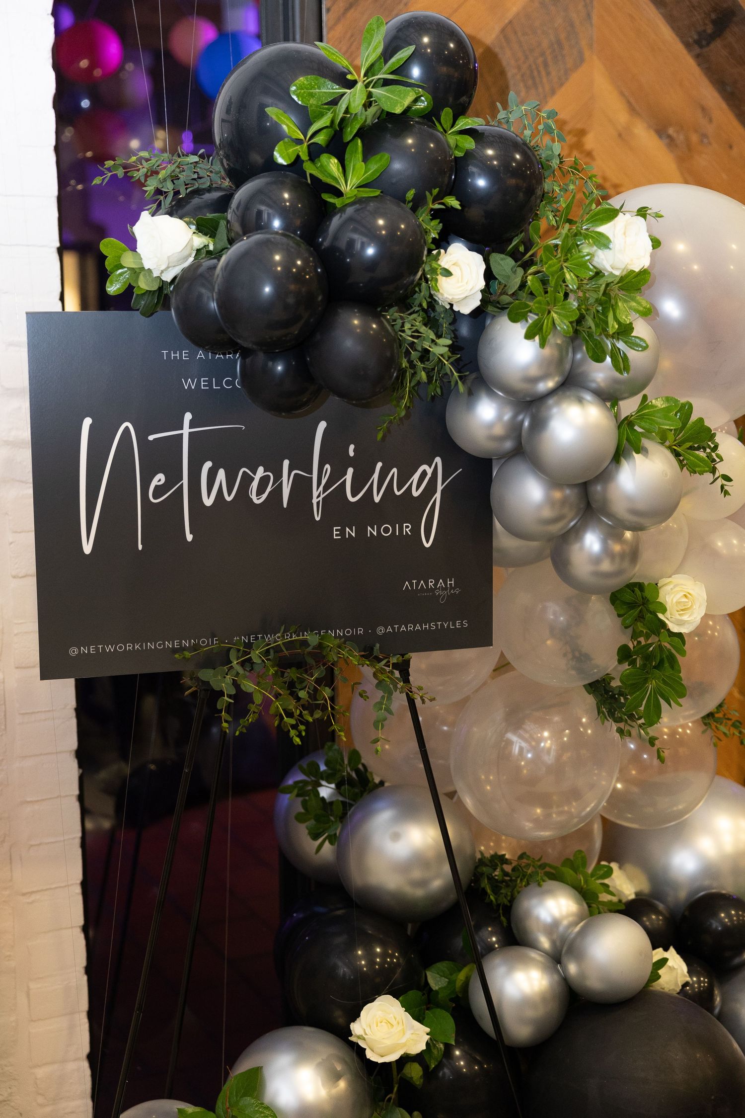 Elegant Networking En Noir welcome sign surrounded by black, silver, and white balloon garland with greenery.