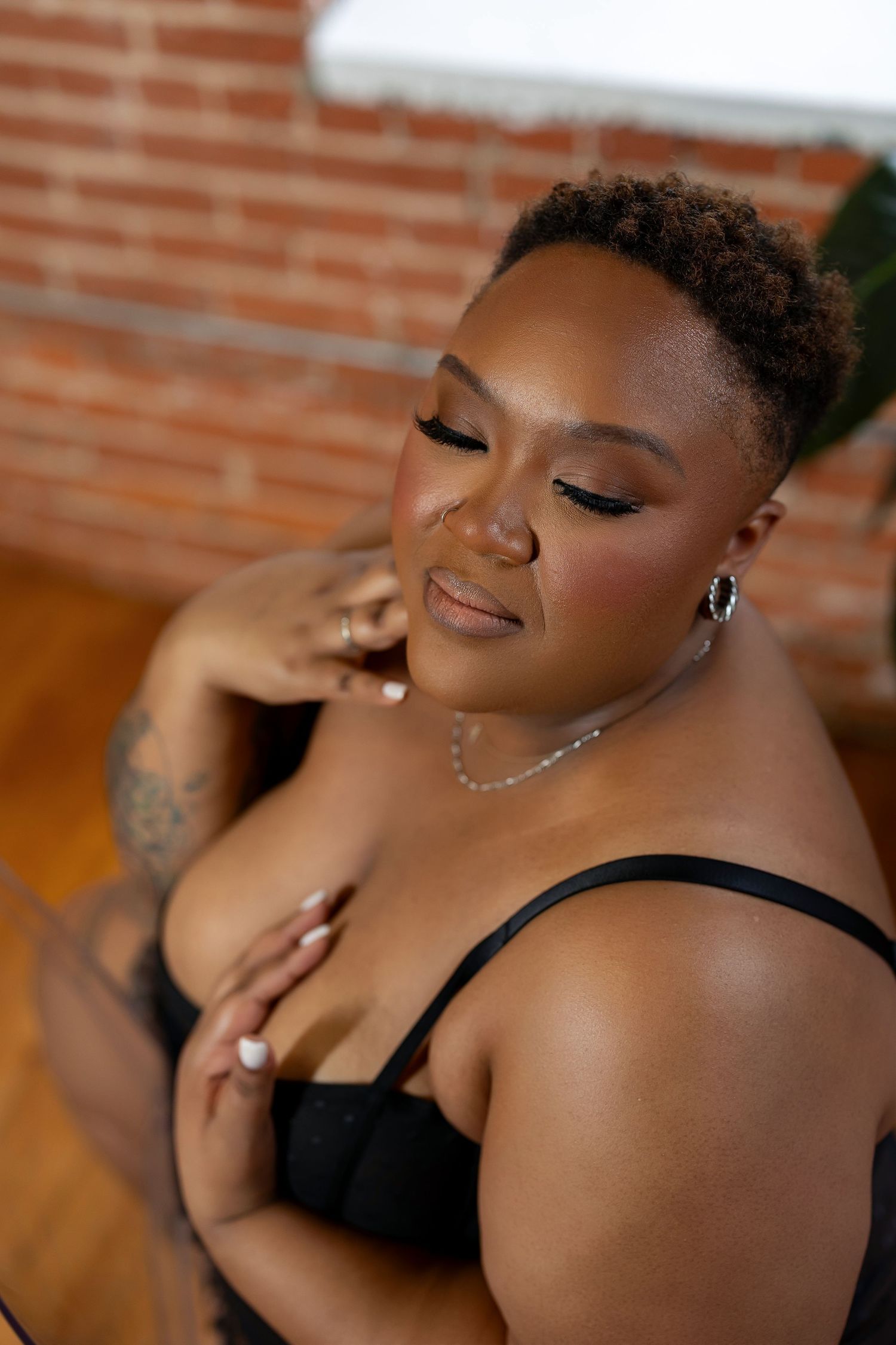A confident plus-size woman in black lingerie poses gracefully in front of a brick wall in a boudoir setting.