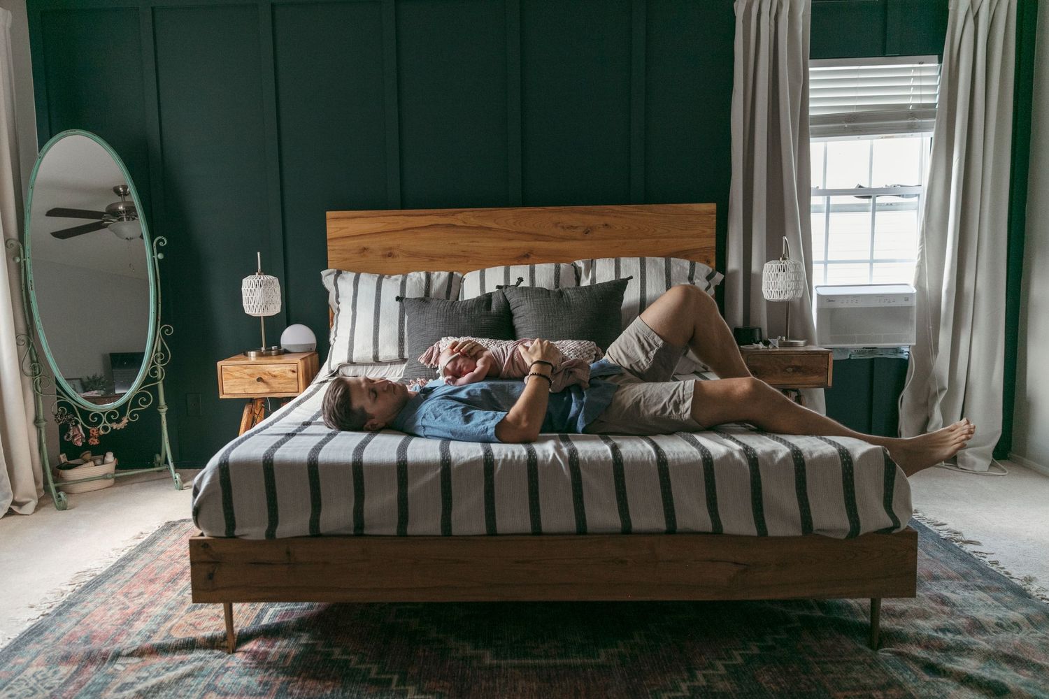 Newborn shoot on modern wooden platform bed with dark green wall and window in background.