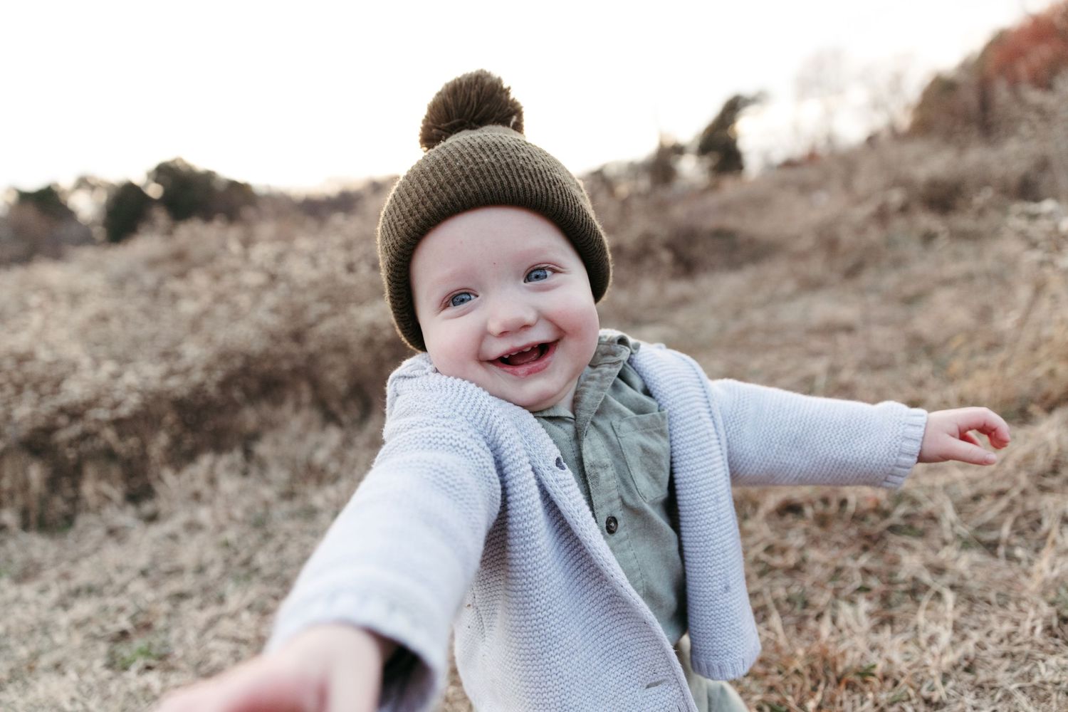 A joyful baby wearing a brown beanie and gray sweater reaches out towards the camera in an outdoor setting.