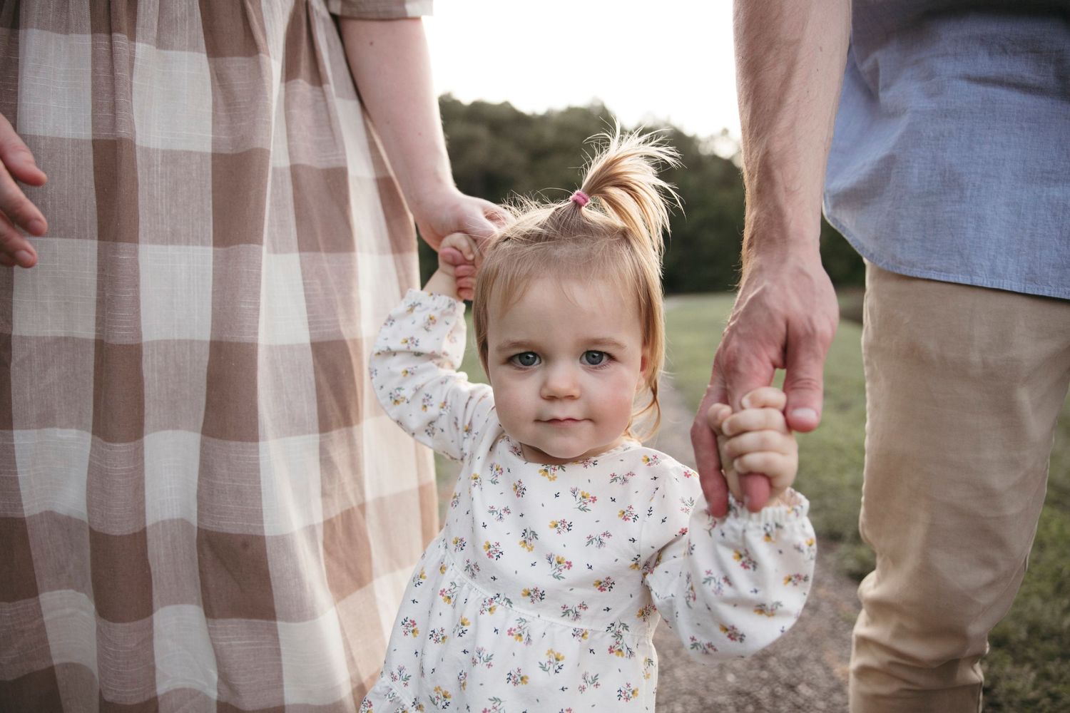 Toddler in plaid dress holding parents' hands while walking outdoors.