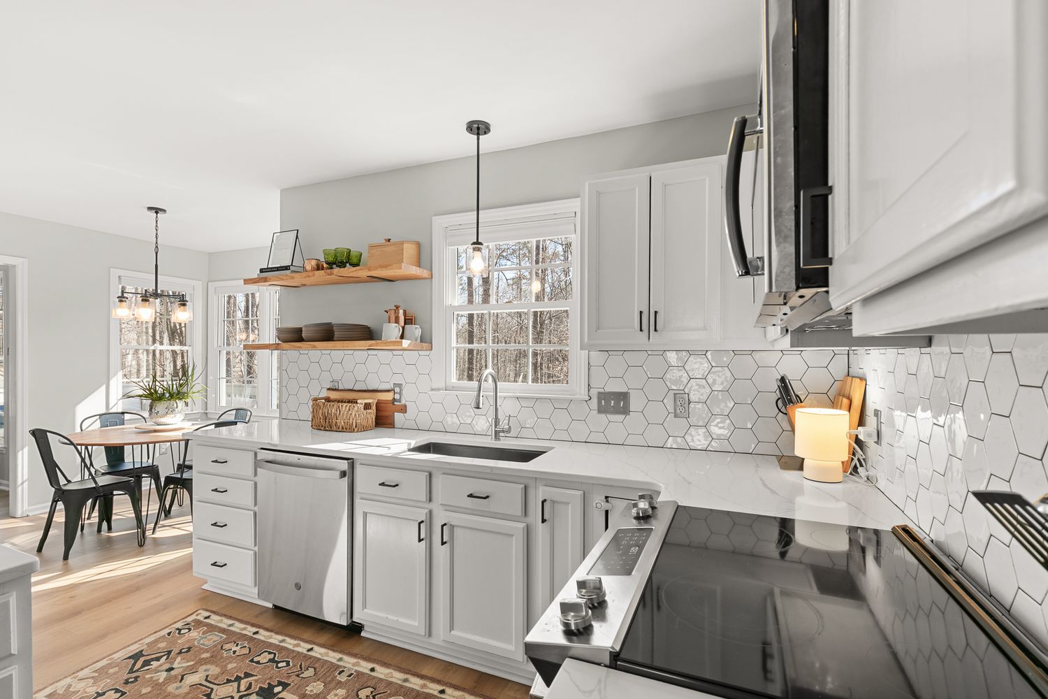 Modern white kitchen with hexagonal tile backsplash, pendant lights, open shelving, and stainless steel appliances.