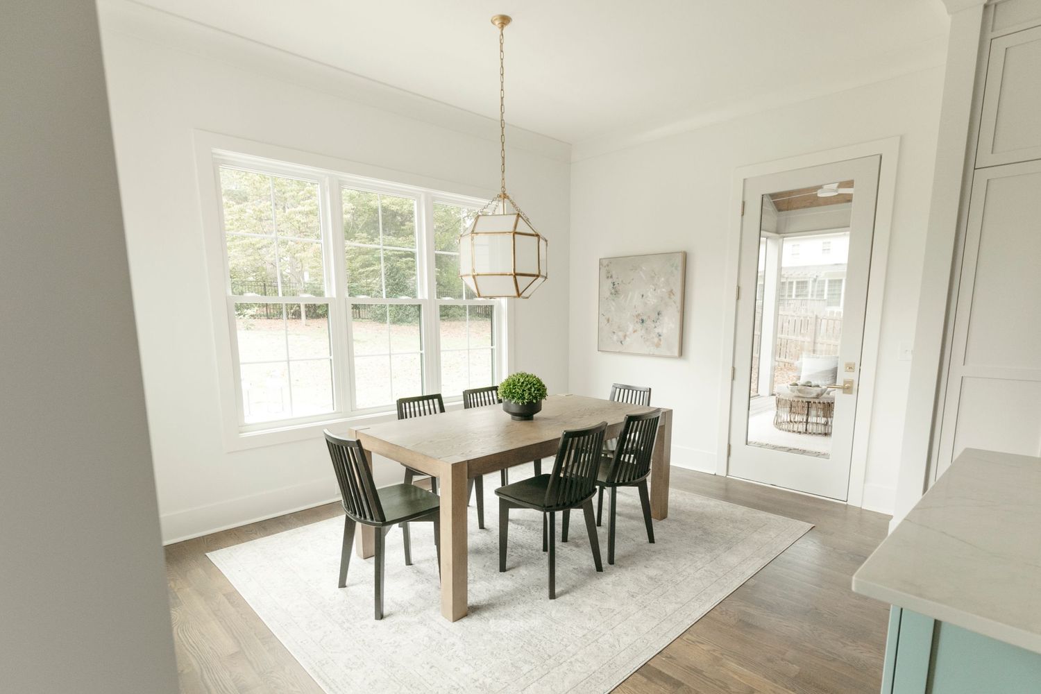 Minimalist dining area with wooden table, black chairs, and bright natural lighting from large windows.
