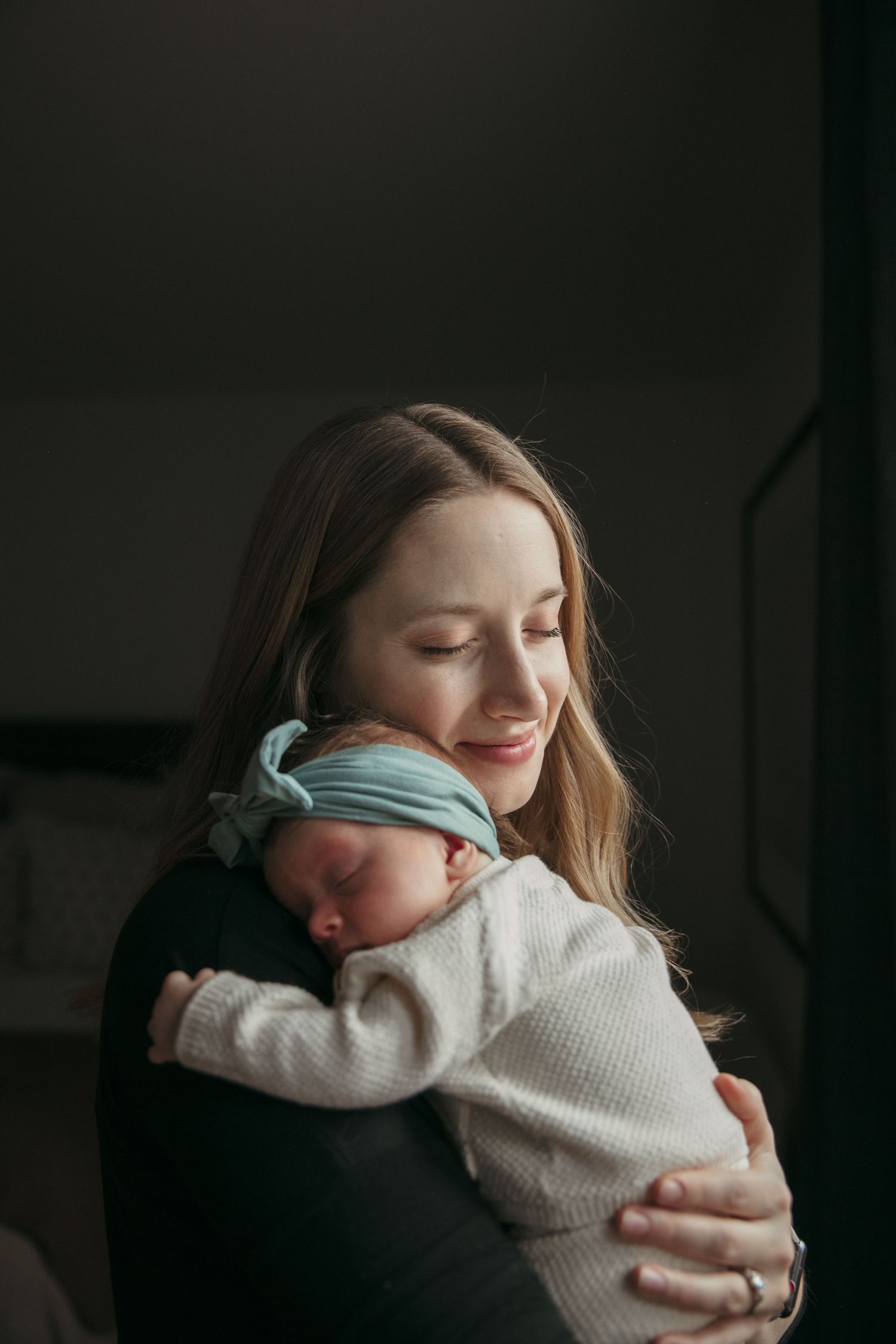 Intimate portrait in moody lighting showing gentle embrace of a newborn in white clothing.