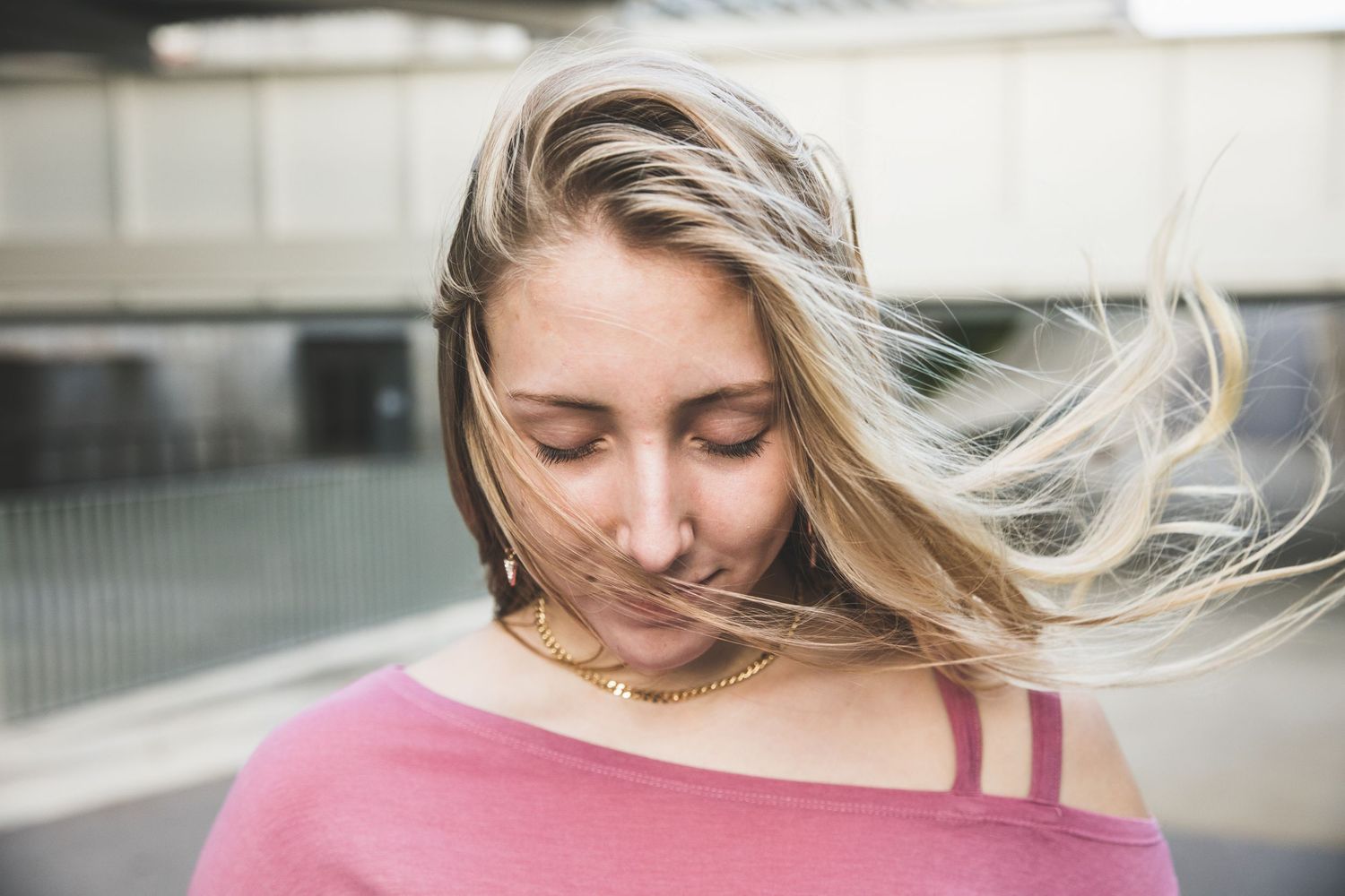 Long blonde hair blows in the wind against an urban backdrop wearing a pink off-shoulder top.