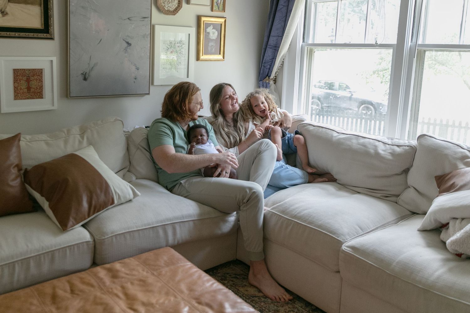 Three people relax together on a comfortable gray sectional sofa by a bright window in a cozy living room.
