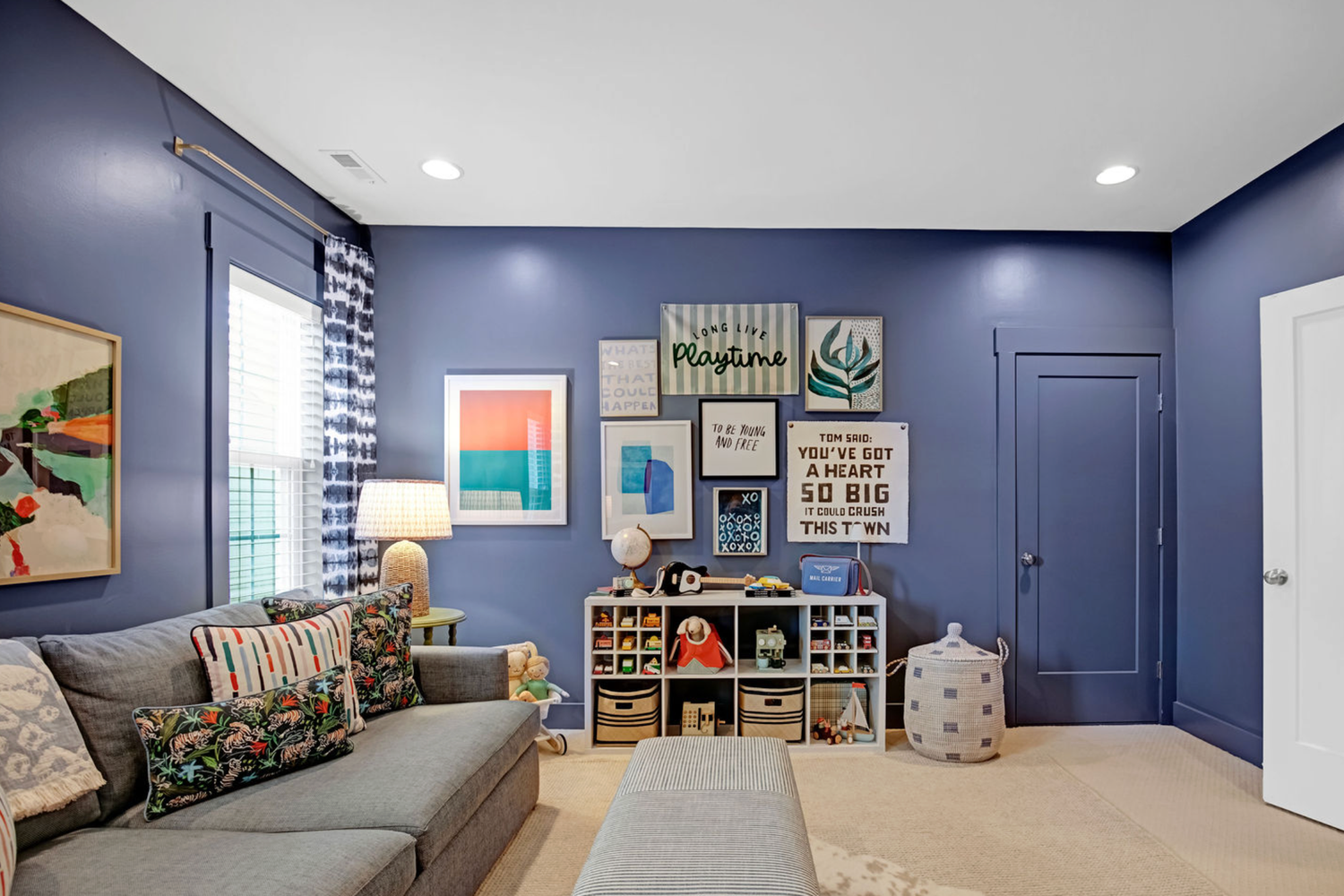 Vibrant blue kids playroom with gallery wall, gray sofa, colorful pillows, toy storage, and light wood floors.