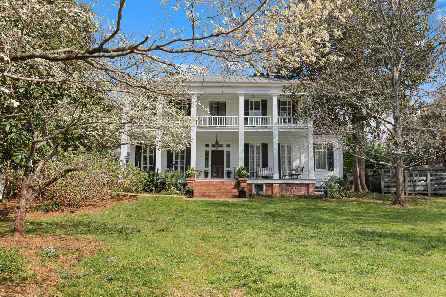 White two-story Southern colonial home with grand columns, wrap-around porch, and lush green lawn surrounded by blooming trees.