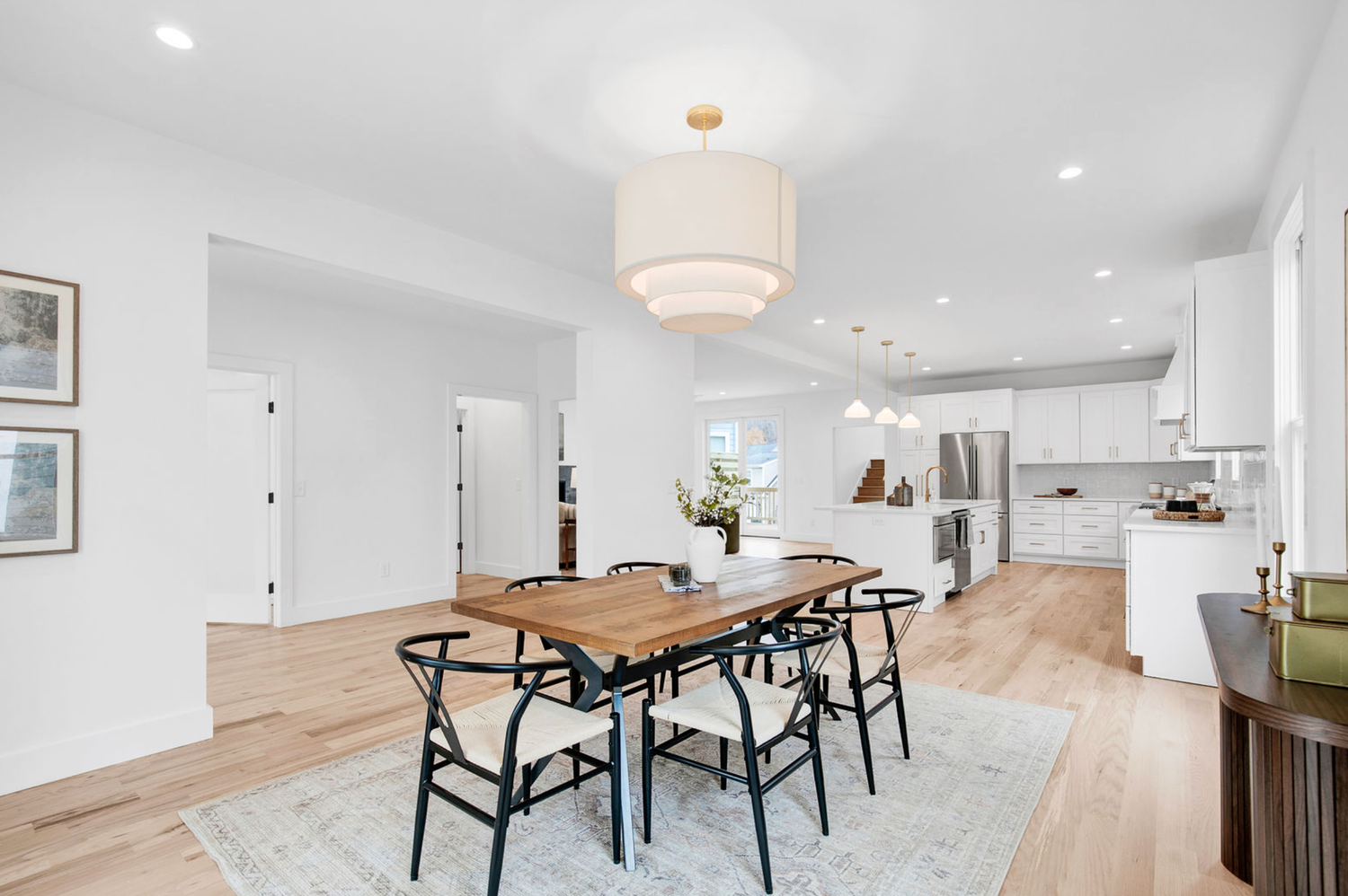Modern open-plan dining room with wooden table, black chairs, pendant light, and white kitchen in background on light hardwood floors.