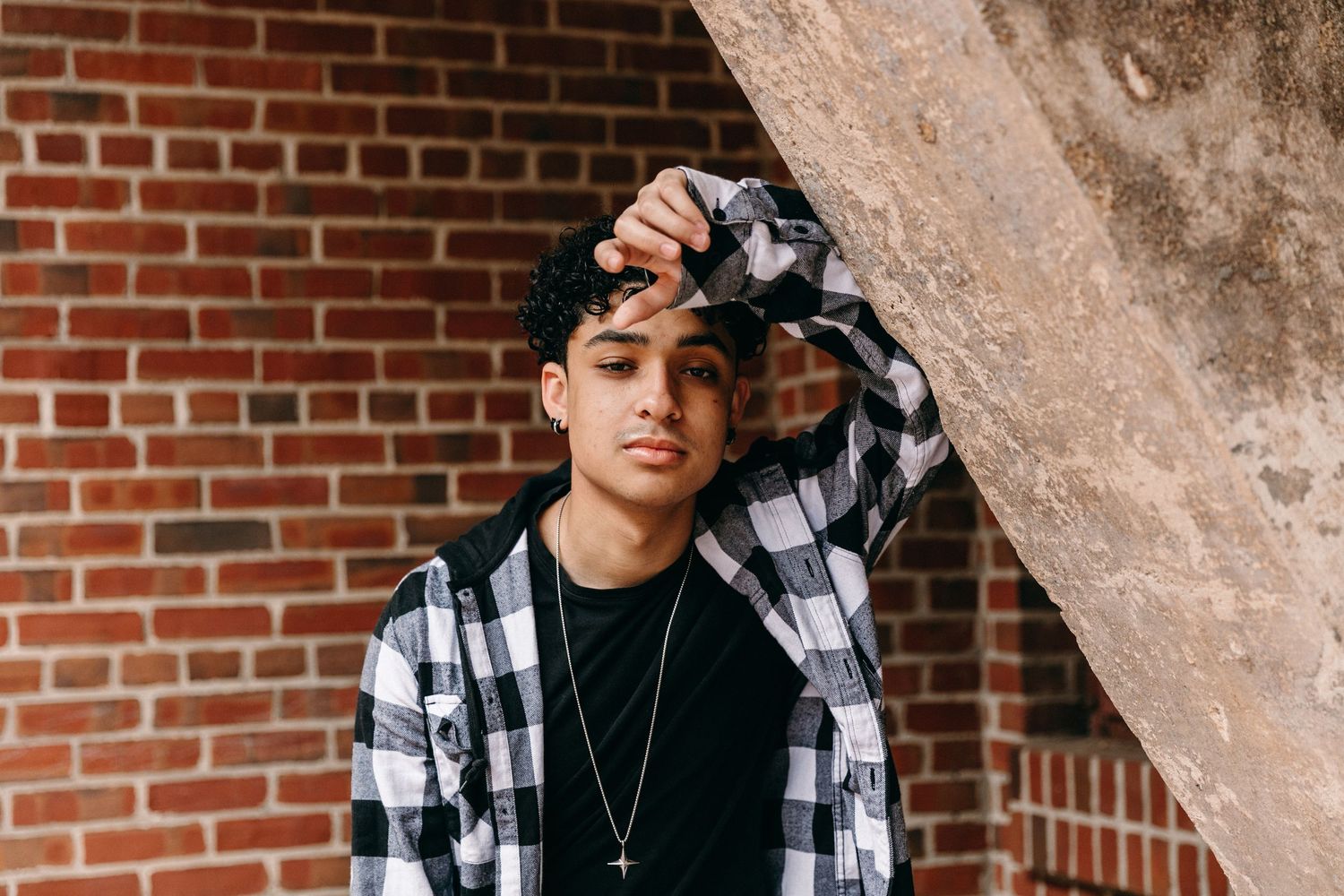 Person in black shirt and plaid overshirt posing near concrete architectural feature.