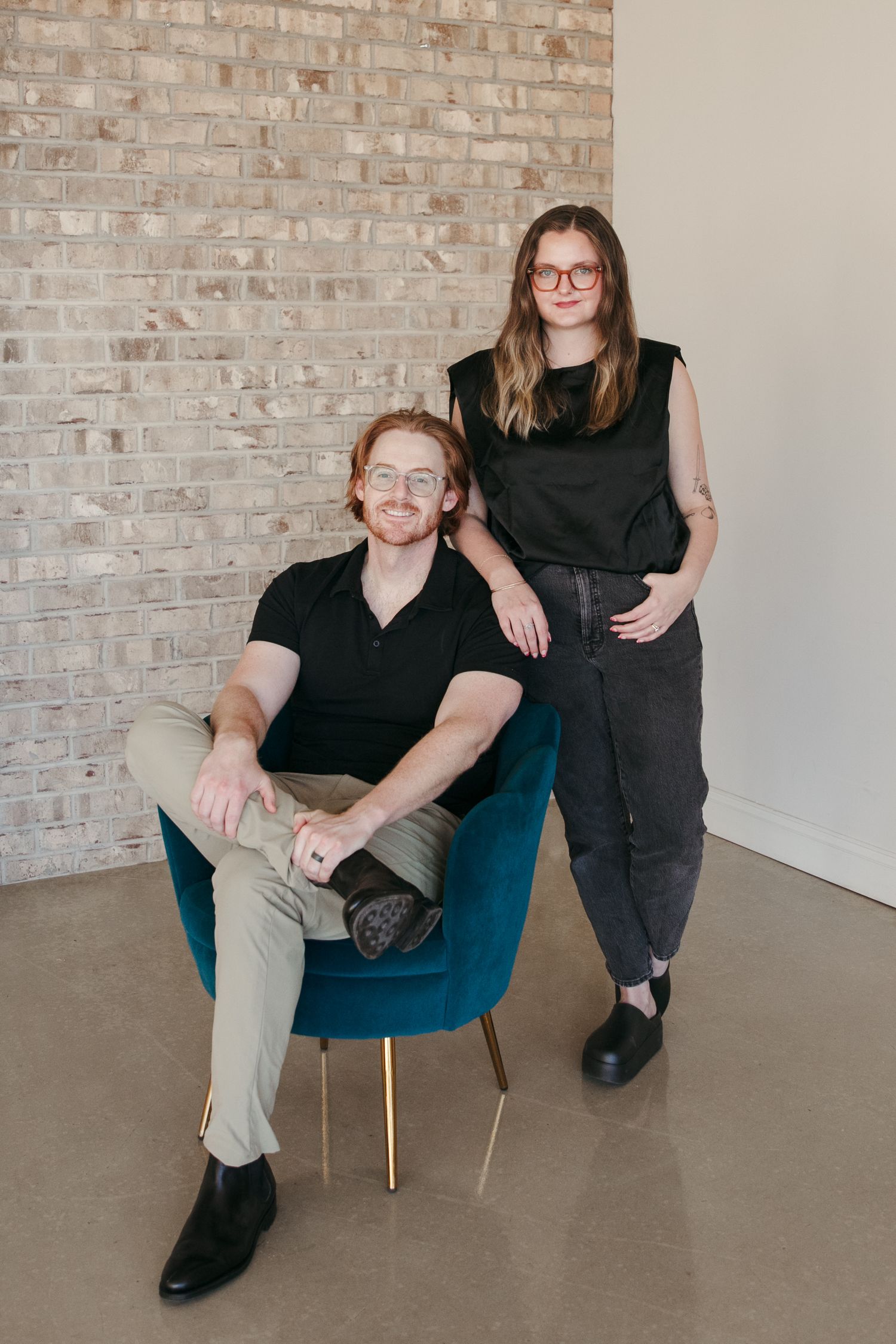 Two people in business casual attire pose against exposed brick wall with modern blue chair.