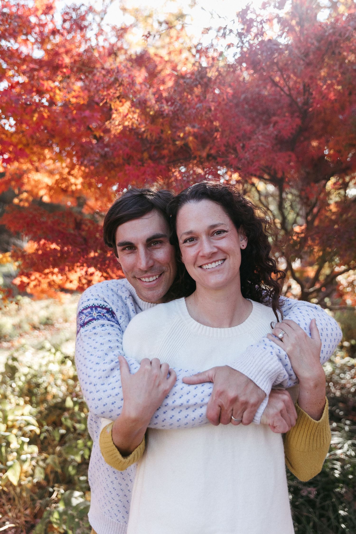 A couple embraces under beautiful red fall trees wearing cozy sweaters.