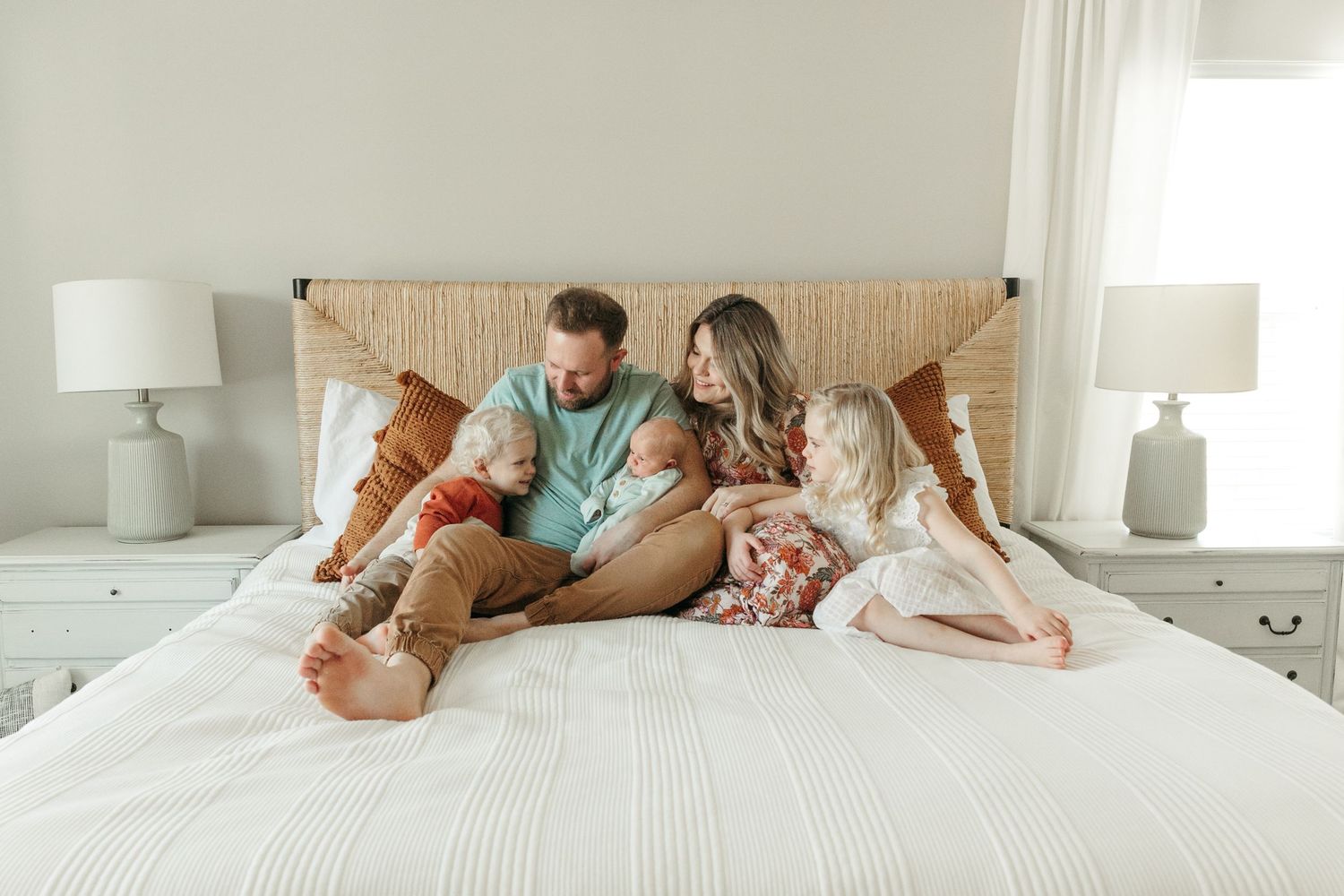 Family relaxing together on a white bed in a bright, sunlit bedroom.