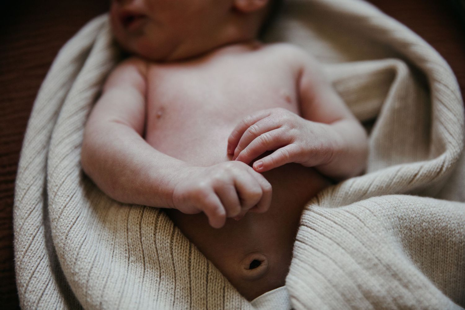 Close-up view of a newborn baby's tiny hands and torso wrapped in a cream-colored knit blanket.