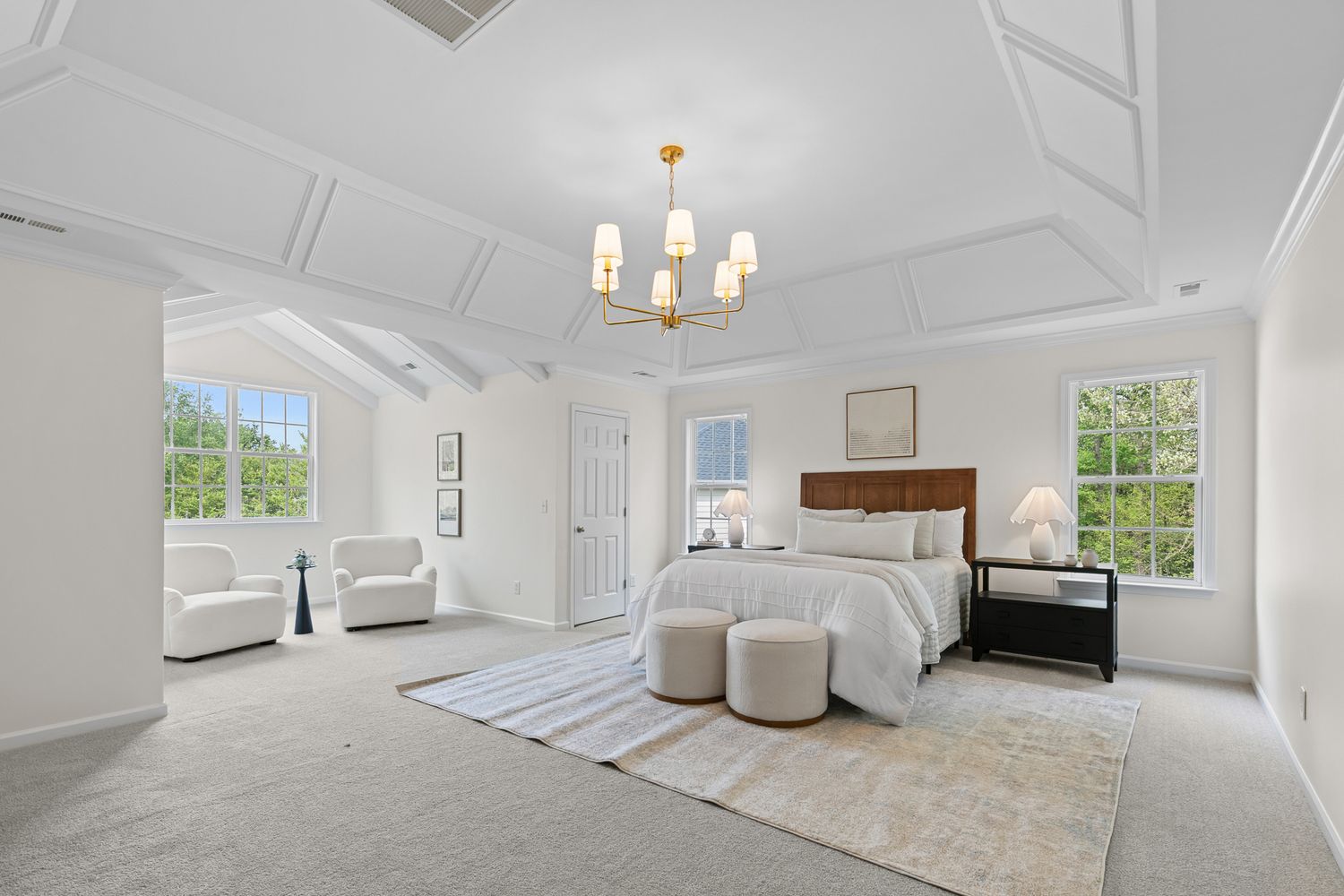 Bright white master bedroom with coffered ceiling, chandelier, neutral carpet, large windows, and elegant wood headboard bed.