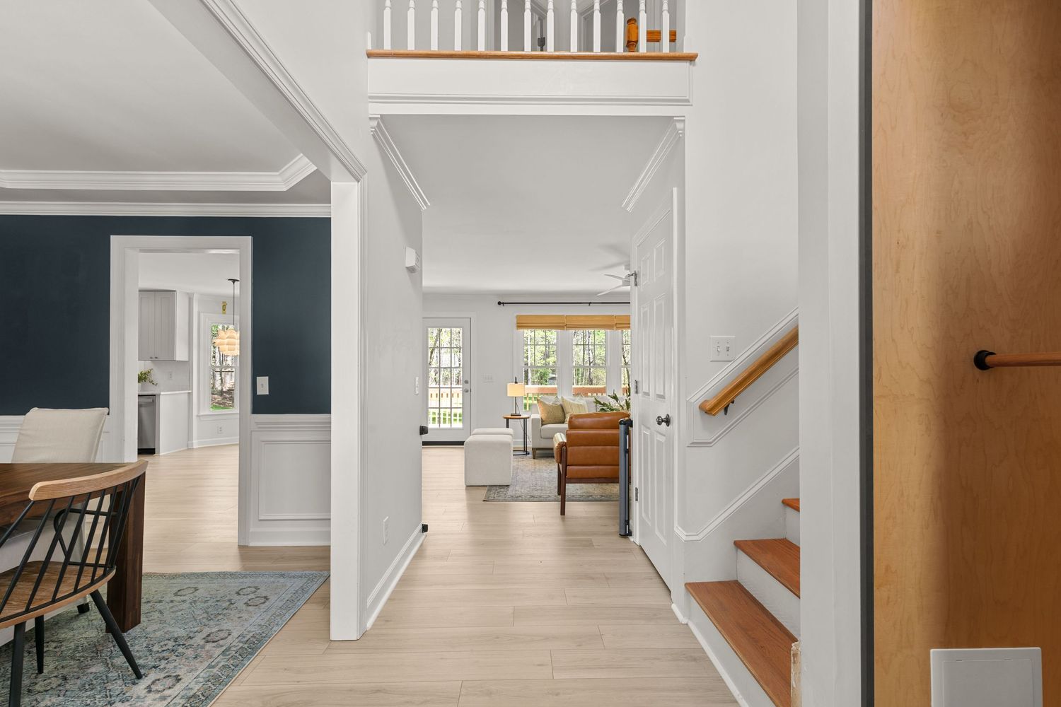 Bright hallway with light hardwood floors, white trim, staircase, and views into a cozy living room and dining area.