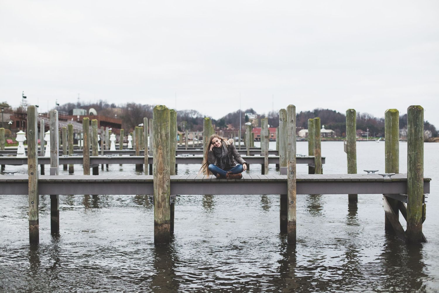 Figure sitting alone on wooden dock overlooking calm harbor waters on cloudy day.