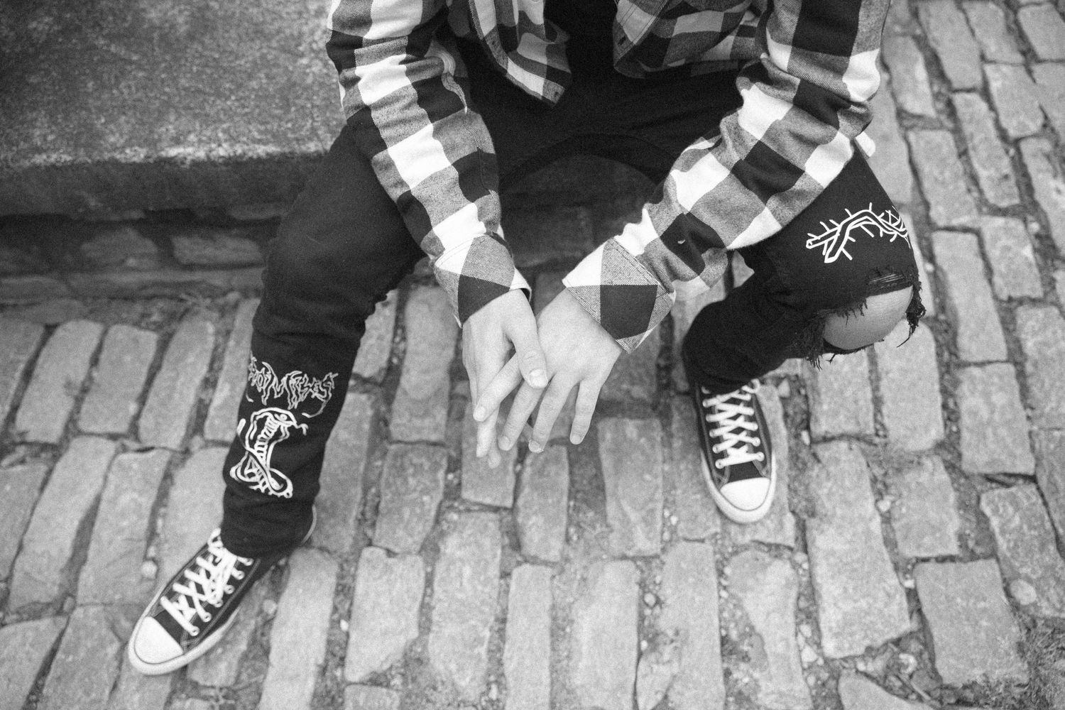 Detail shot of distressed black jeans and classic black and white sneakers on brick pavement.