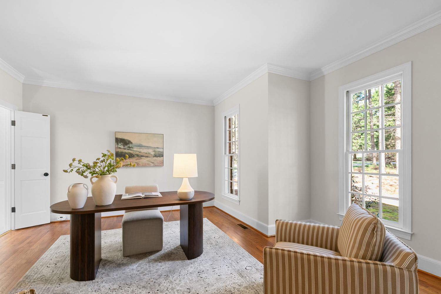 Bright home office with hardwood floors, oval wood desk, striped armchair, and large window overlooking greenery.