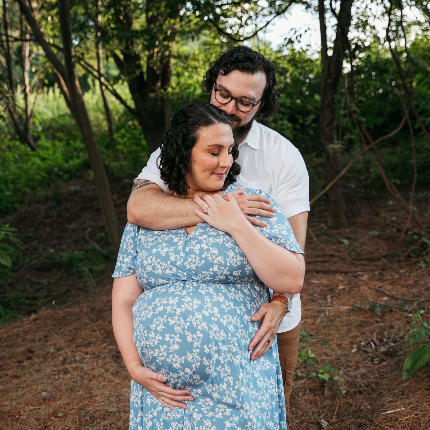 An expectant couple shares an intimate embrace in a wooded setting.