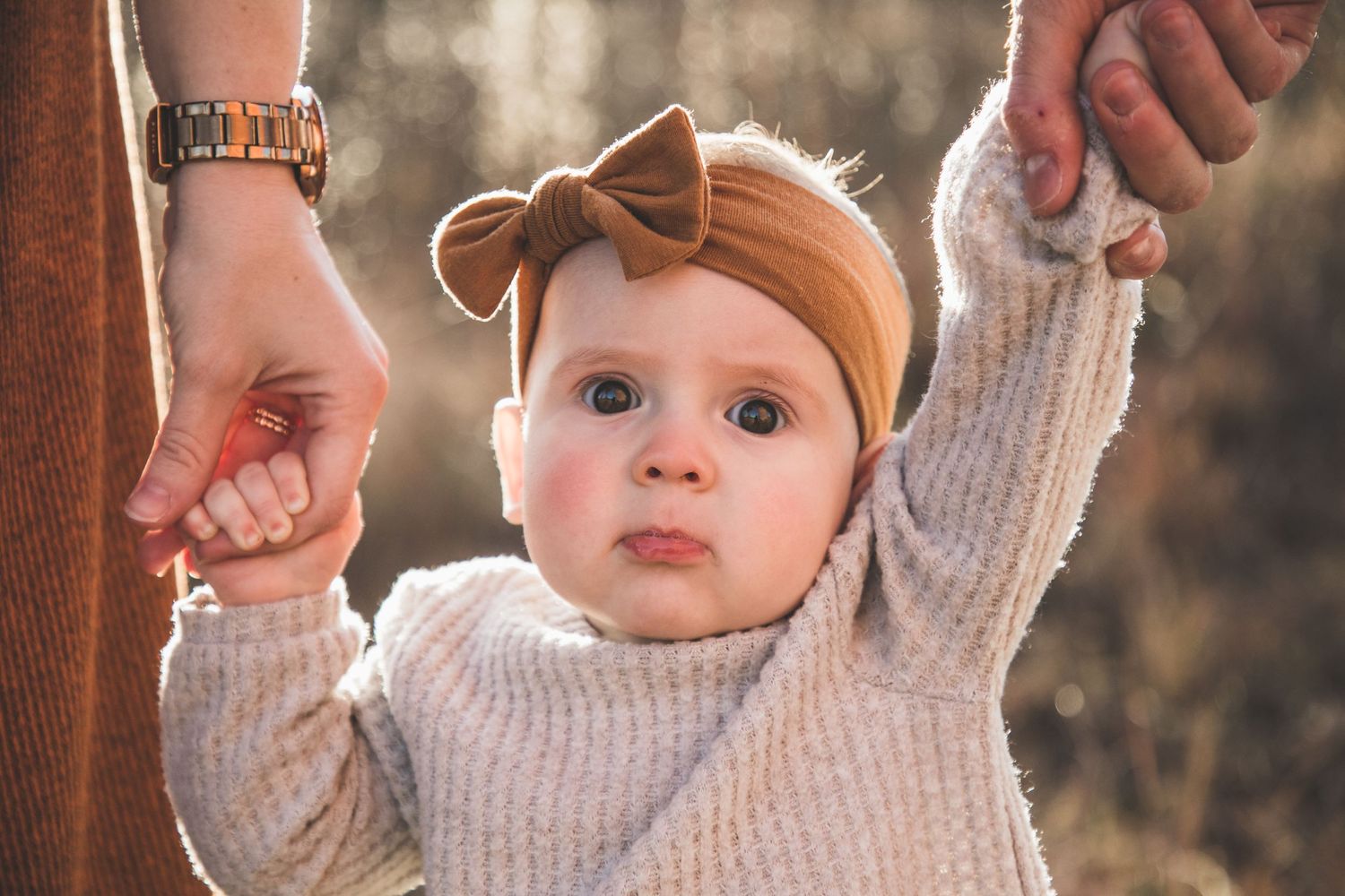Baby in a knit sweater wearing a brown headband reaches up while being supported by hands.