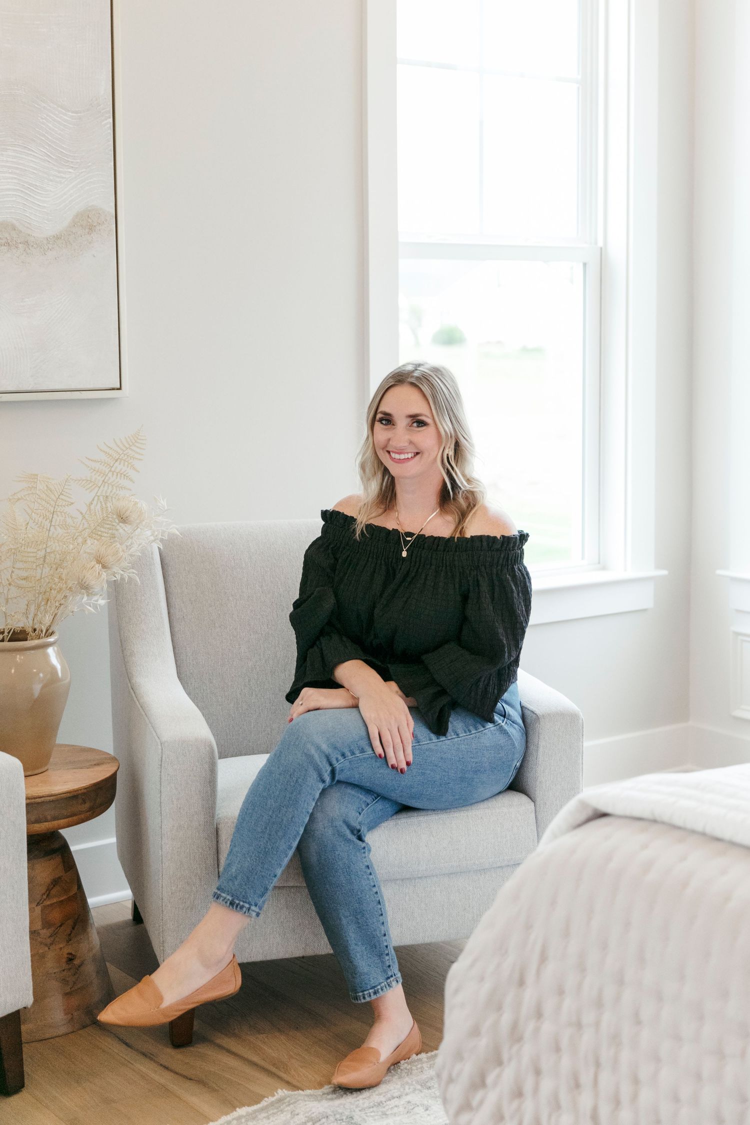 Casual indoor portrait on light gray armchair wearing black peasant top and distressed jeans.
