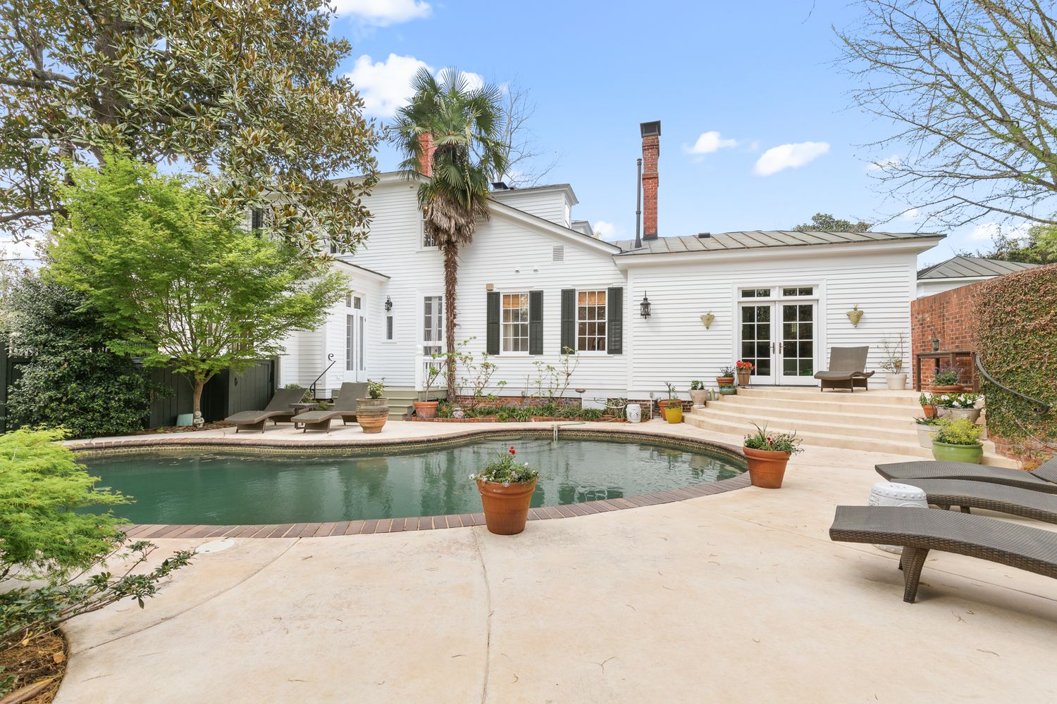 White two-story home with oval pool, stone patio, lounge chairs, and lush landscaping on a sunny day.