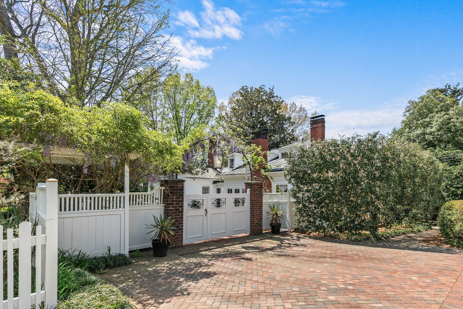 White picket fence and brick driveway lead to a charming white cottage surrounded by lush mature trees and greenery.