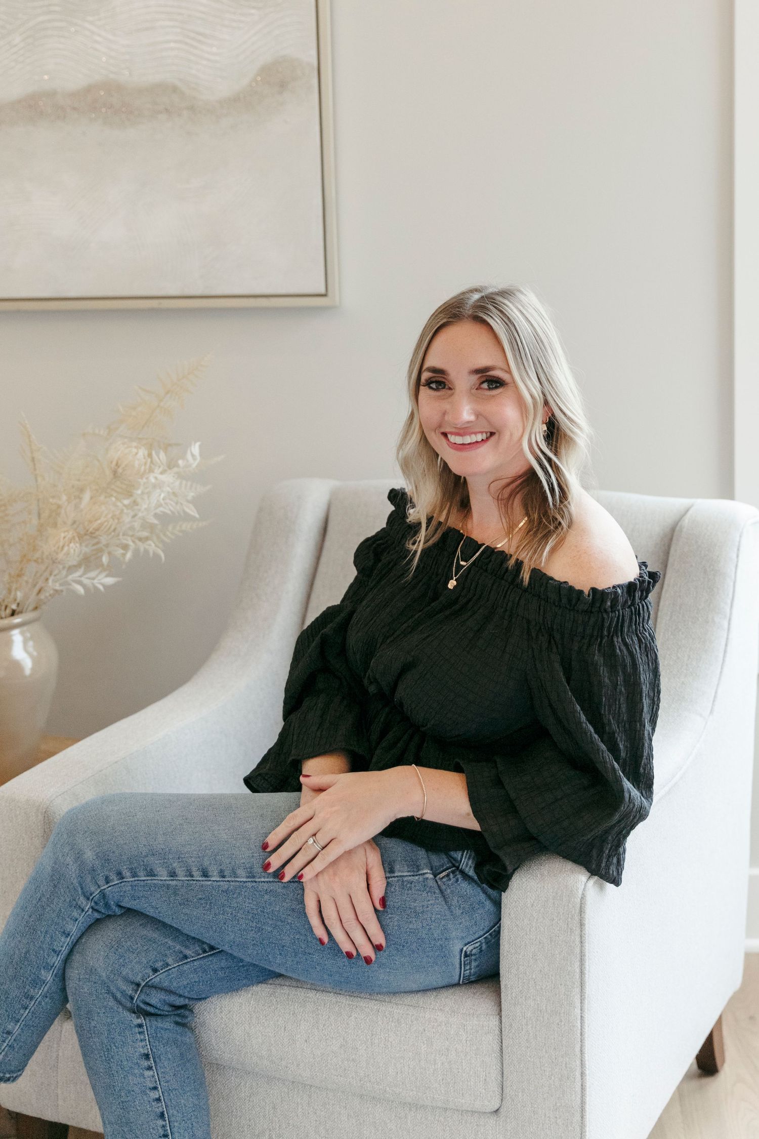 Casual portrait in black off-shoulder top and jeans against neutral home decor background.