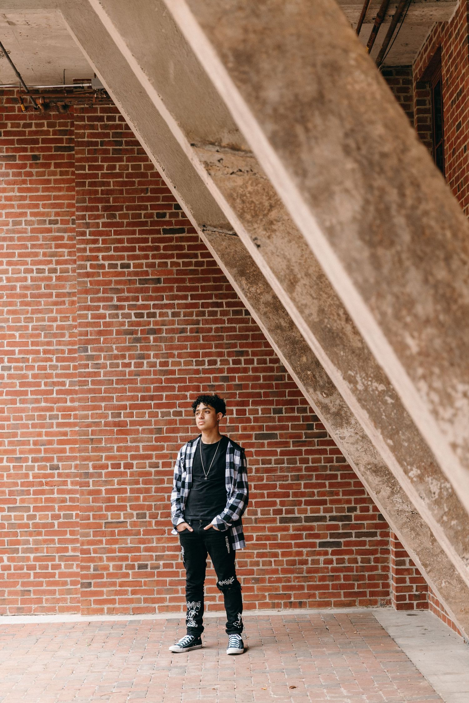 Person in plaid shirt stands under concrete stairway against brick wall.