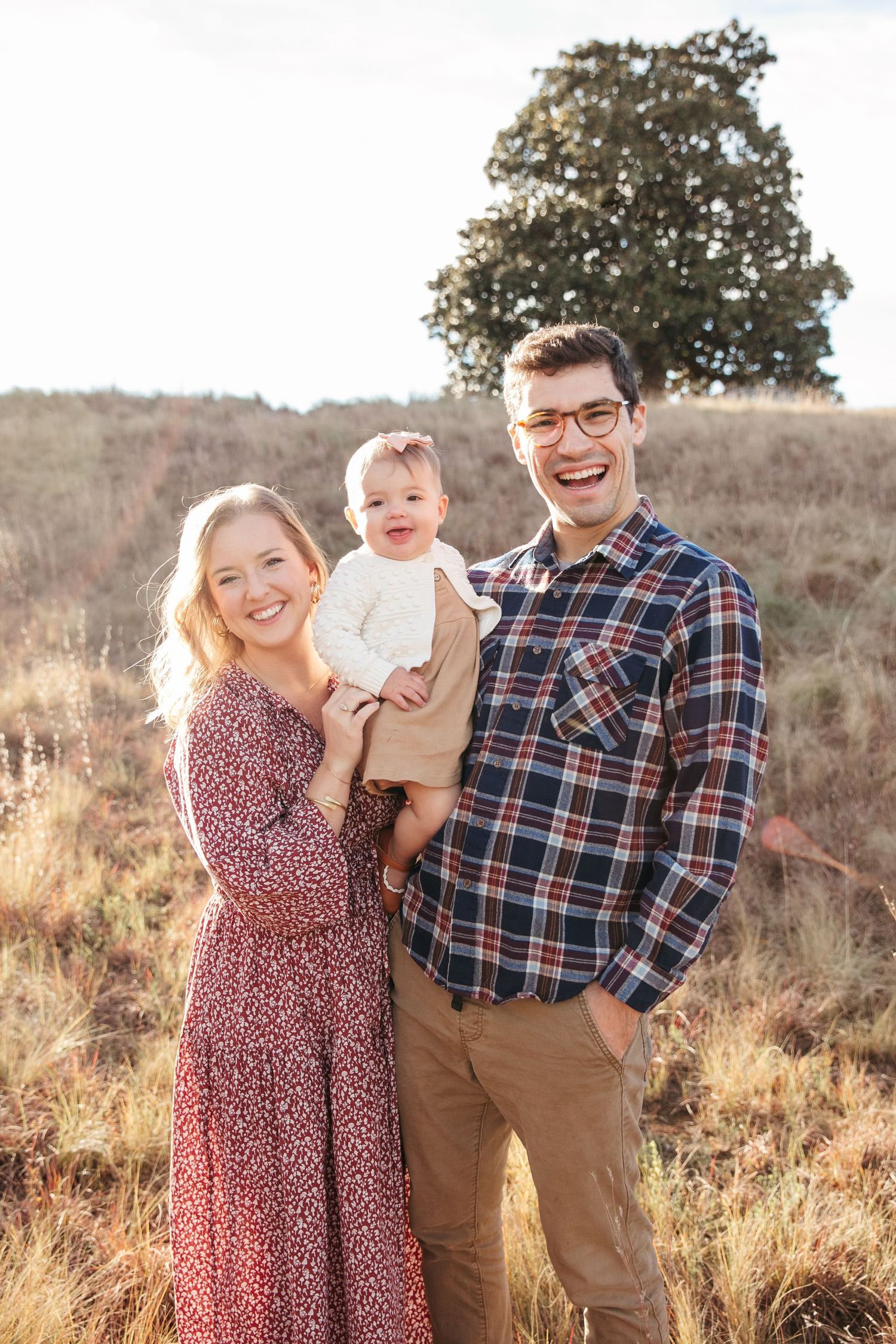 A couple and their baby pose in a golden field at sunset with a tree in the background.