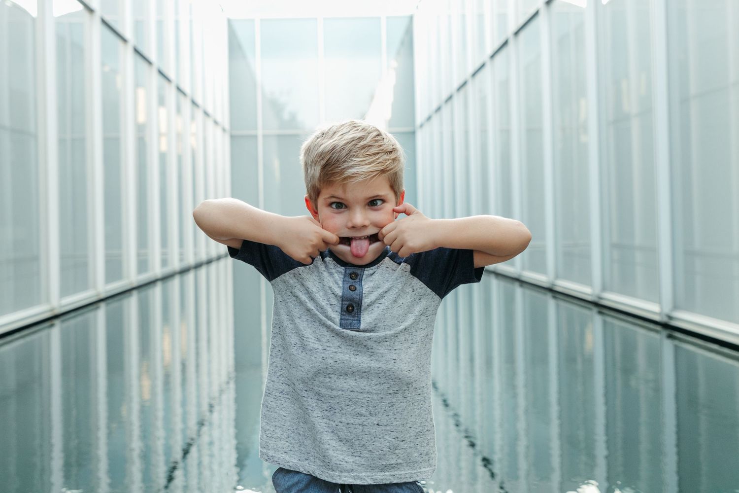 Young child making silly face with hands behind head in industrial corridor.
