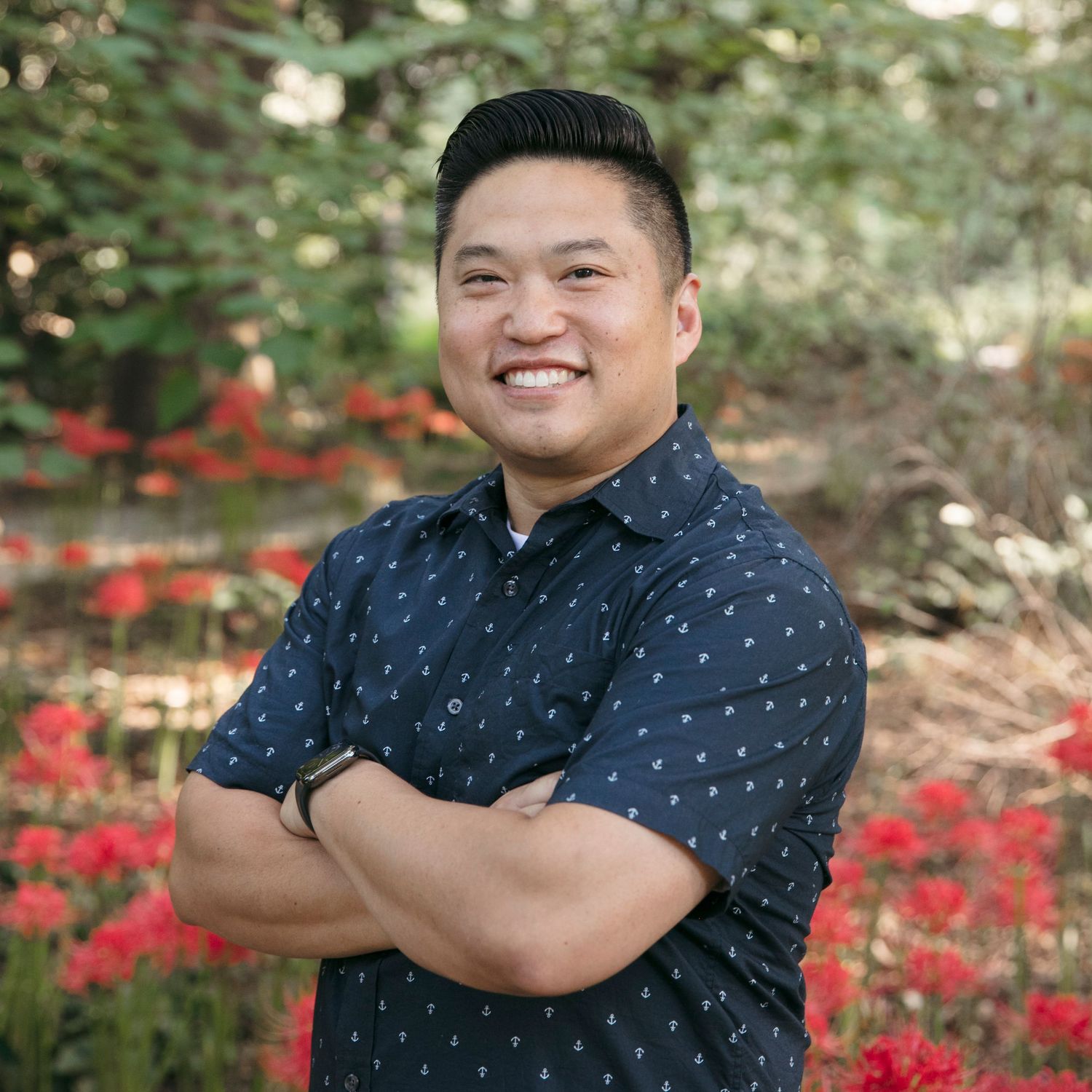 A person in a polka dot navy shirt smiles with crossed arms in front of red flowers and greenery.