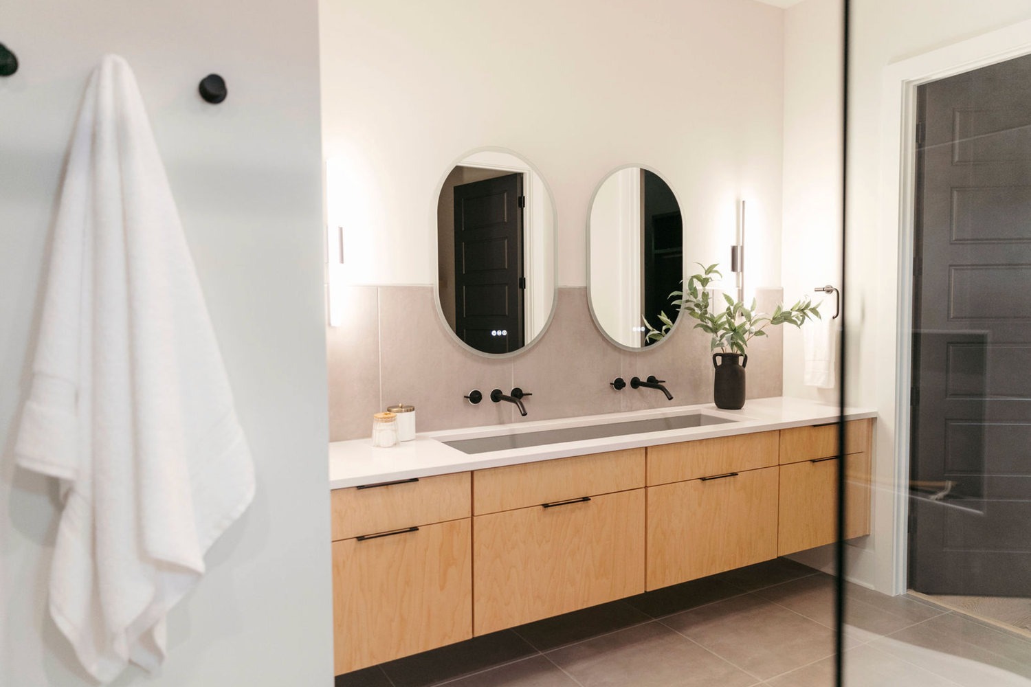 Modern floating double vanity with wood cabinets, dual oval mirrors, and white countertop in a bright minimalist bathroom.