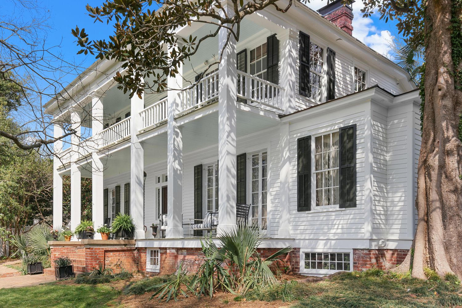 Historic white antebellum plantation home with tall columns, double porches, black shutters, and brick foundation surrounded by trees.