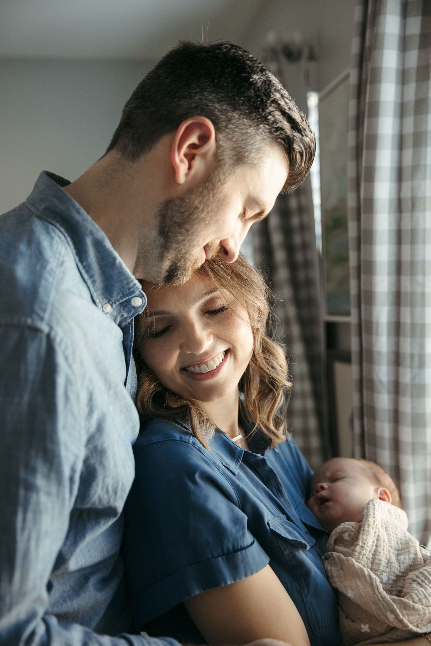 A tender family moment as parents share an intimate moment with their sleeping newborn by a window.