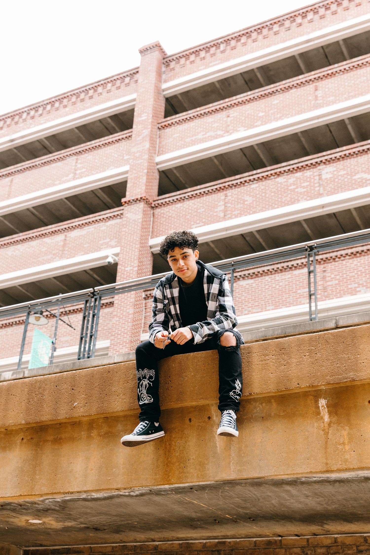 Person in plaid shirt and jeans sits on concrete ledge against brick building facade.