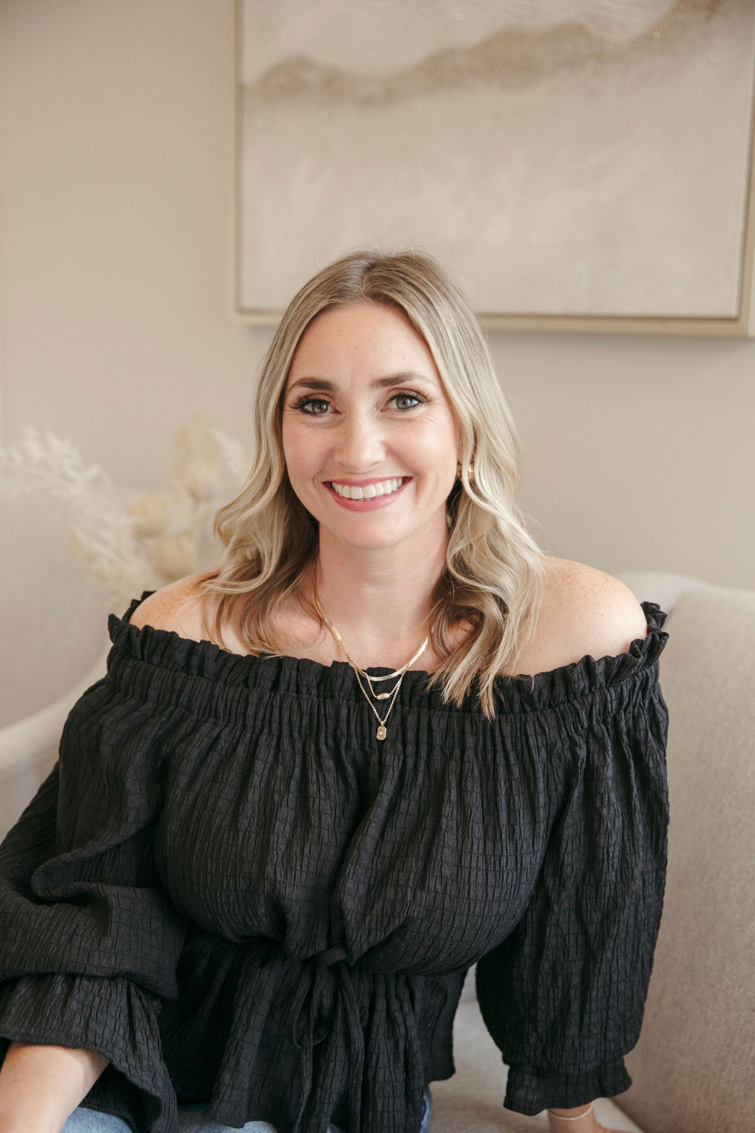 Lifestyle portrait in black off-shoulder top with neutral home decor and art in background.