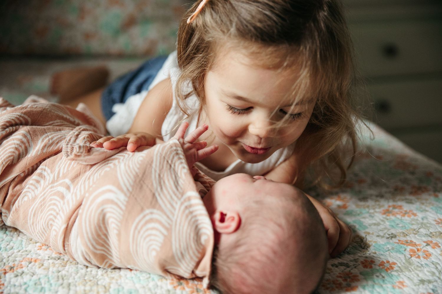 Gentle sibling interaction with newborn on floral patterned bedding.