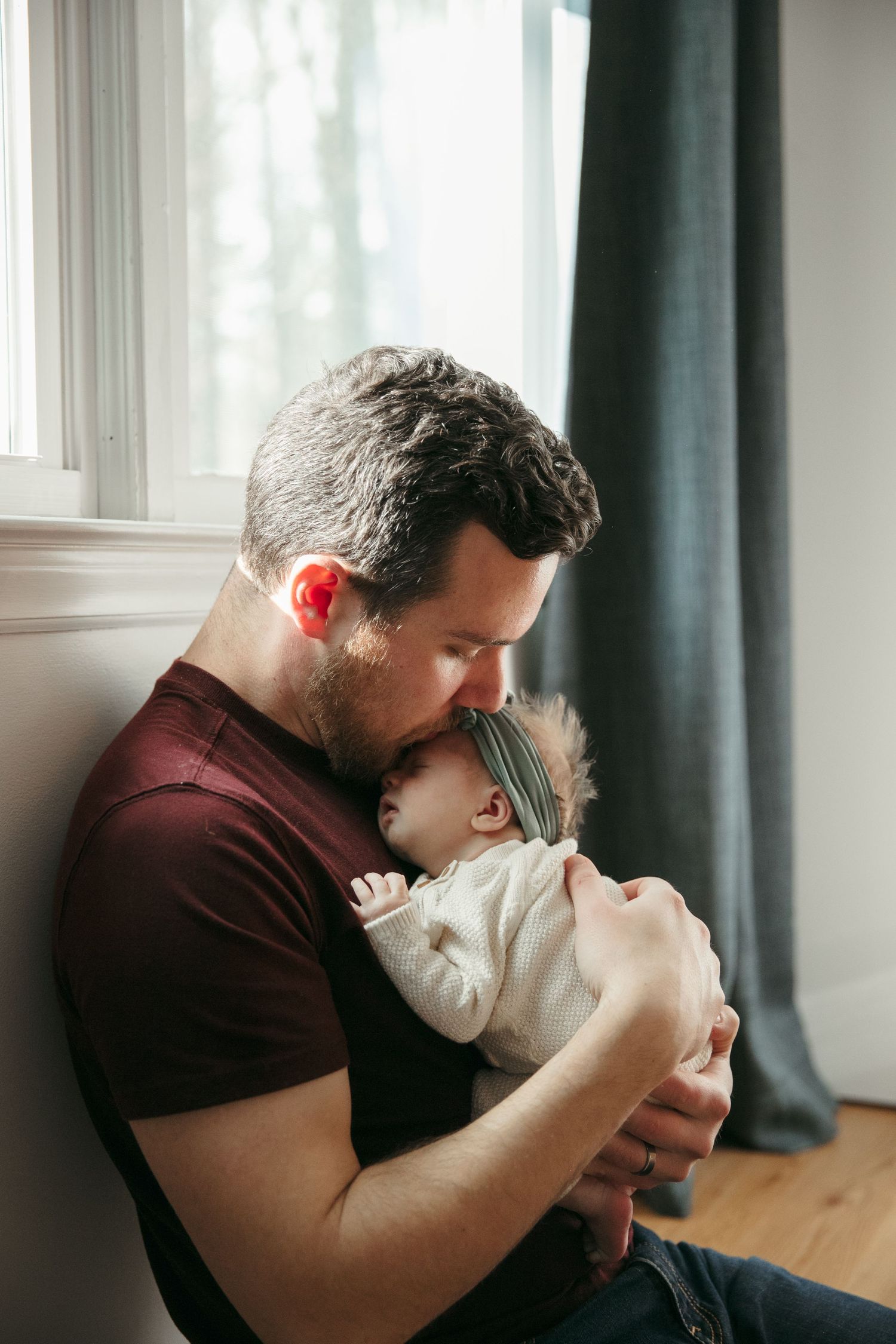 A tender moment between parent and infant by a window with dark curtains in a cozy room.