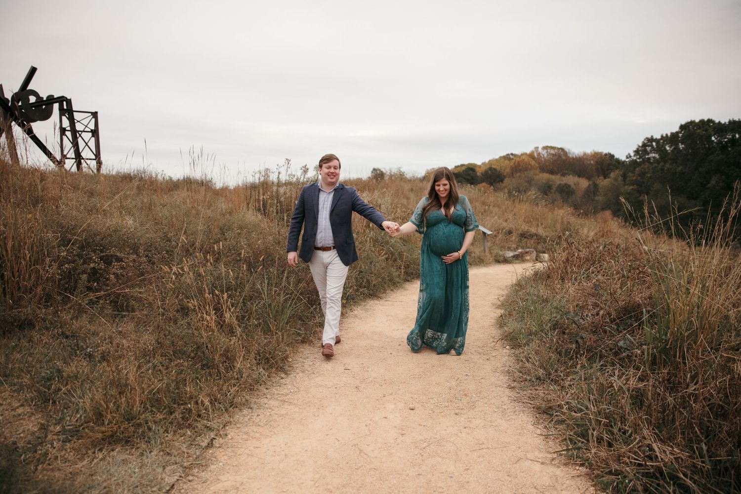 Pregnant couple walking along dirt path through golden field during sunset photoshoot.
