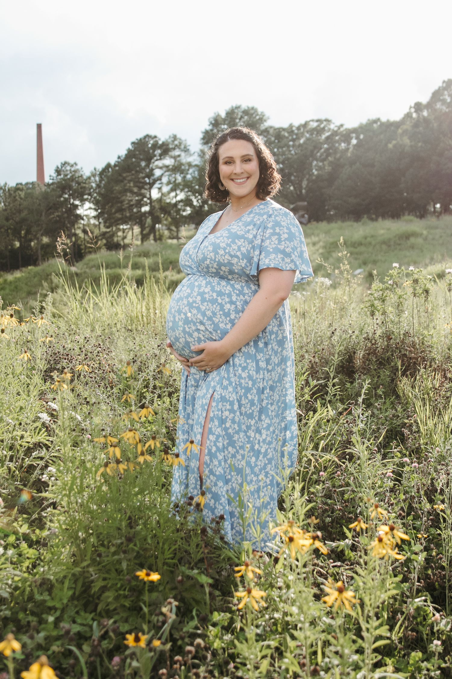 Expectant mother in blue floral dress poses in wildflower field during golden hour.