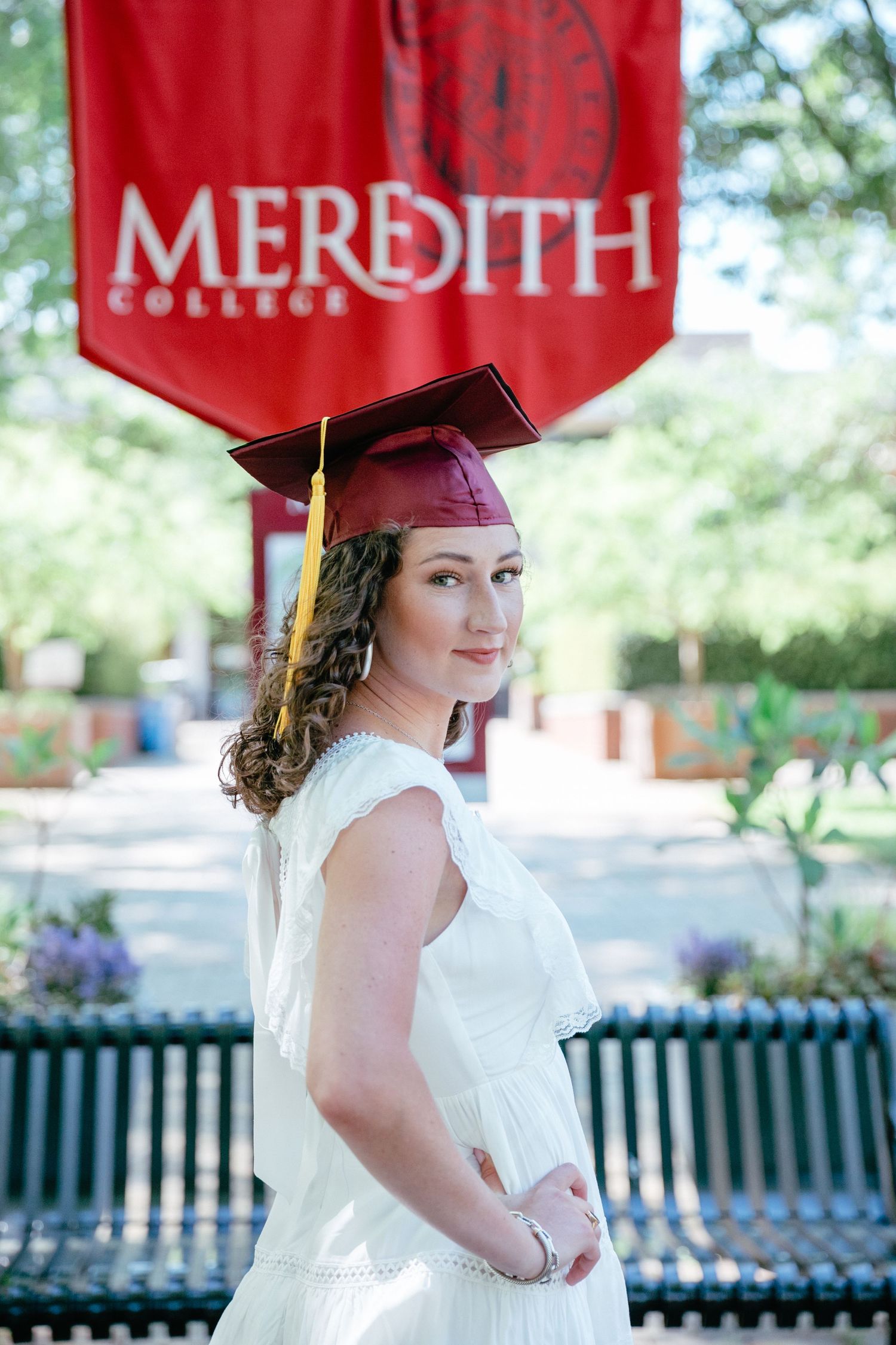 Graduate in red cap and white dress poses outdoors on university campus.