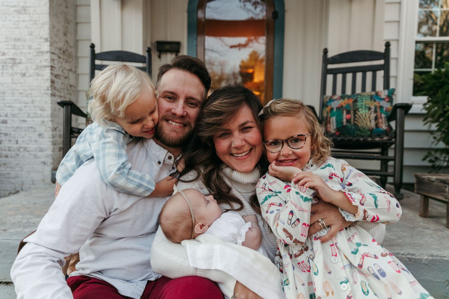 A family group sits together on a front porch with white brick and fall decorations.