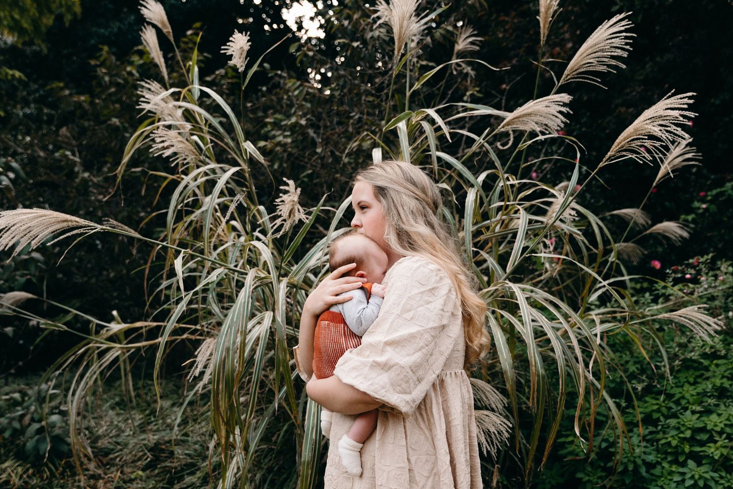 A person in a cozy beige sweater stands against tall pampas grass plumes in a moody outdoor setting.