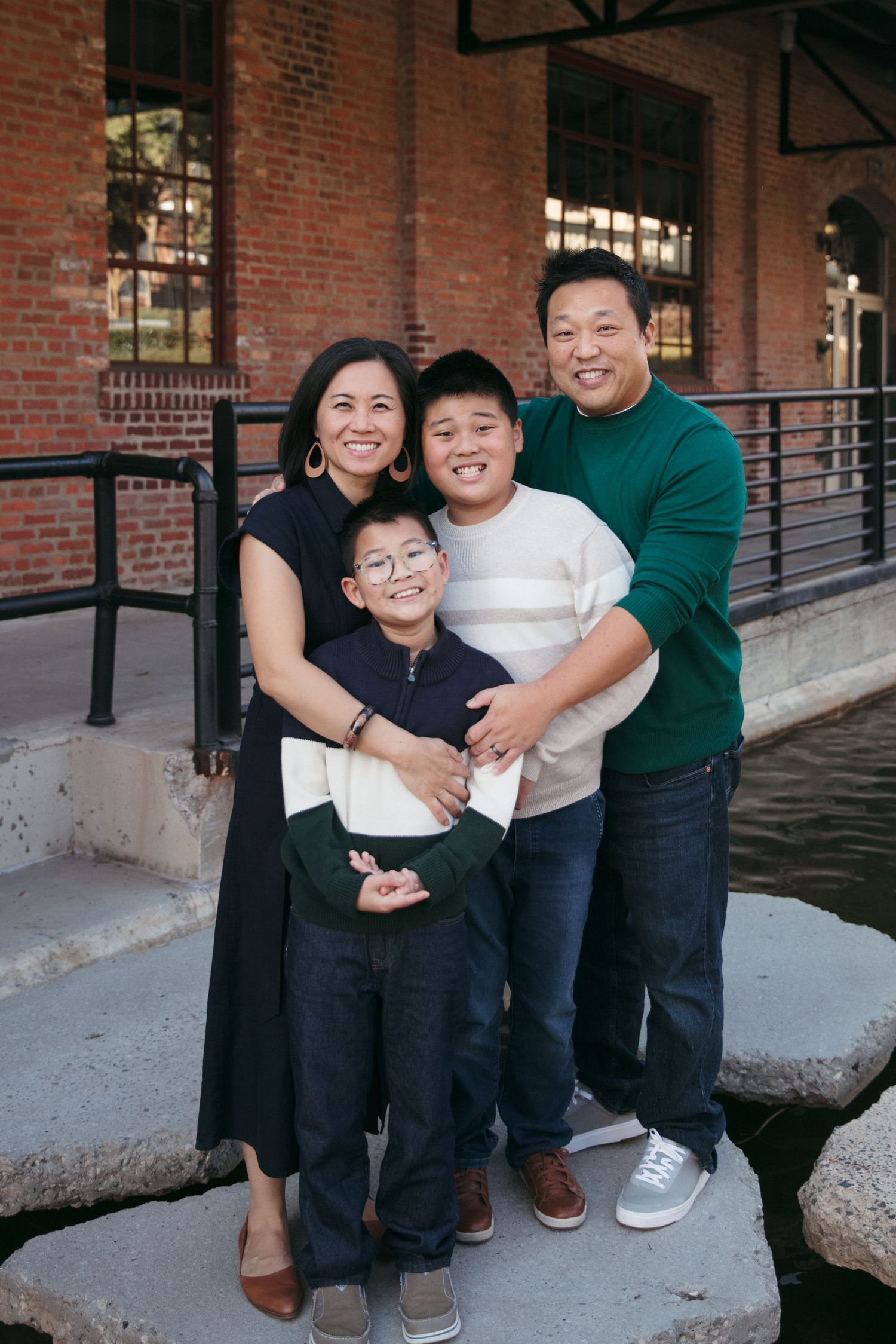A joyful family of four poses together outside a brick building with industrial architecture.