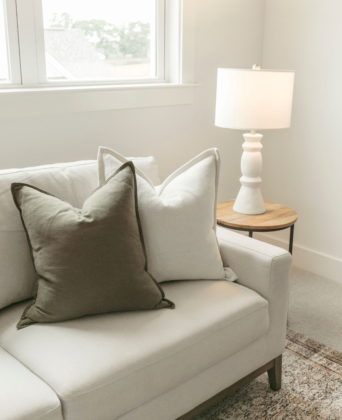 White sofa with olive and cream throw pillows beside a wooden side table with a white lamp near a bright window.